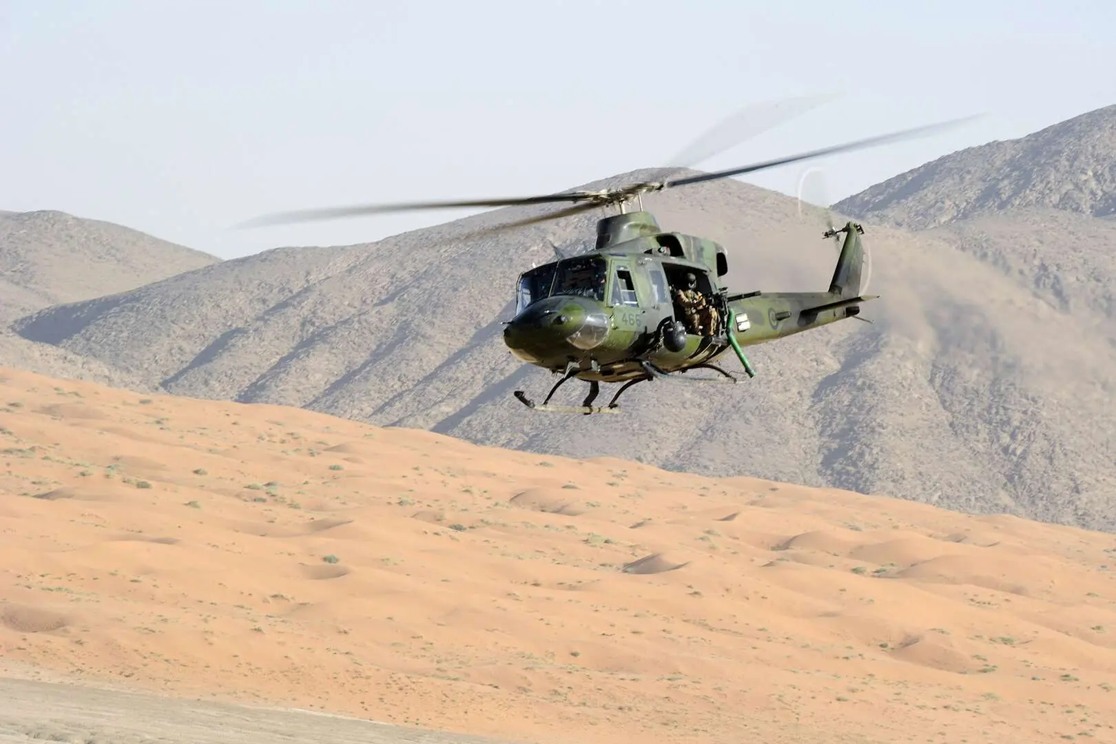 Un hélicoptère vert des Forces armées canadiennes survole un décor désertique et montagneux. La porte de côté de l’appareil est ouverte et on peut apercevoir un mitrailleur assis, tenant son arme entre ses mains.