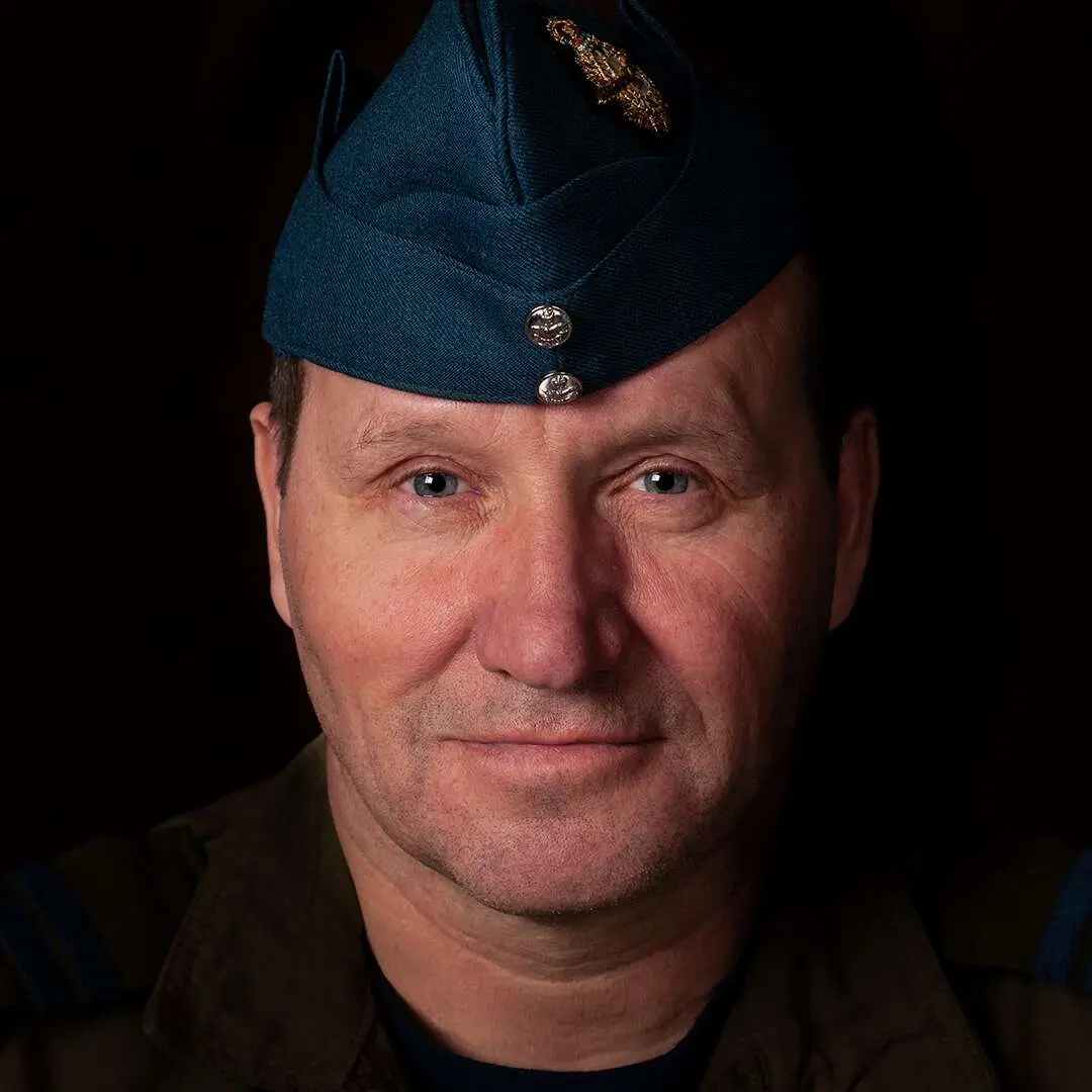 Sur un fond noir, le visage du capitaine Gérard Caron portant le calot bleu roi de l’Aviation royale canadienne. Il regarde la caméra avec un léger sourire.