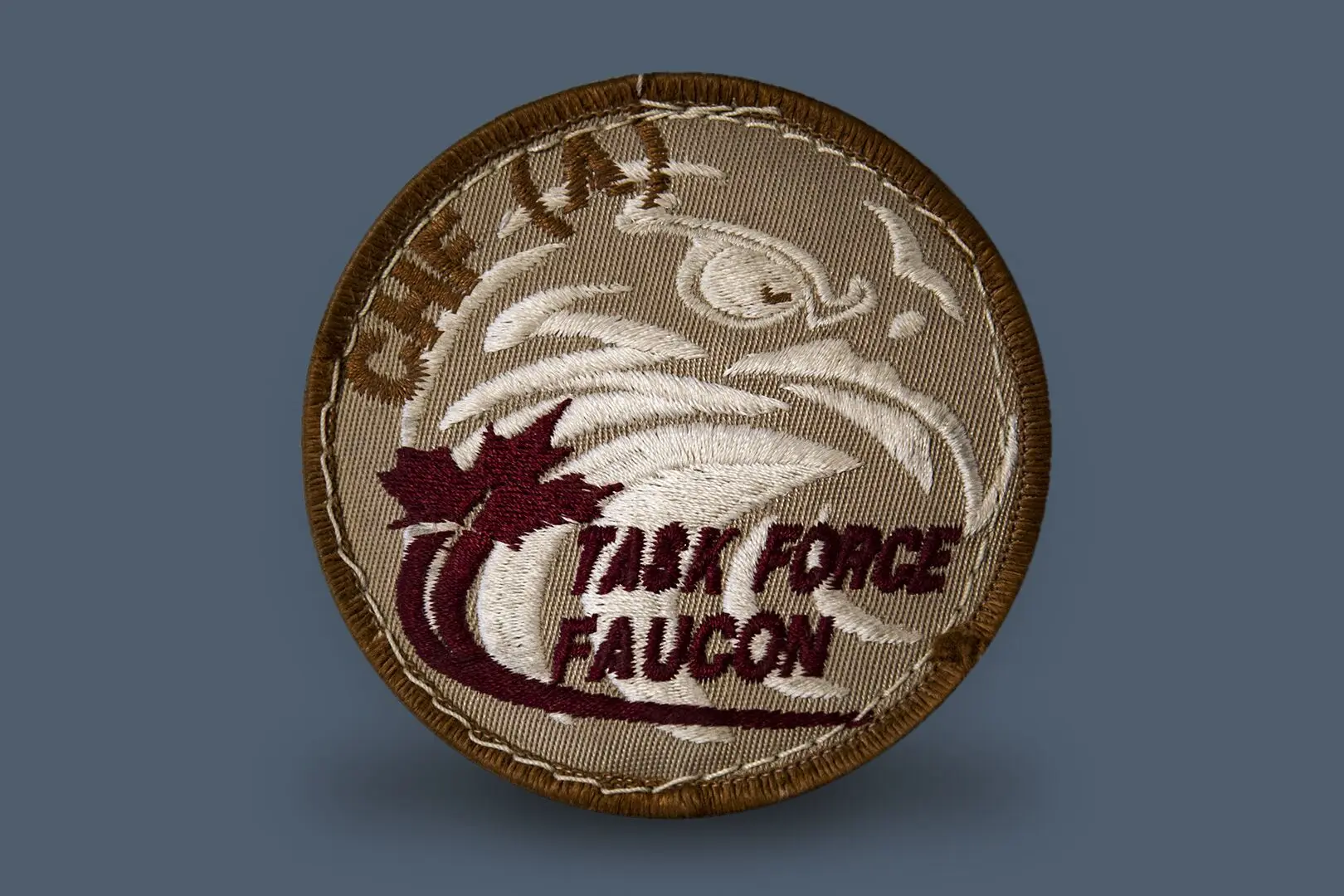 Écusson de couleur beige avec un profil blanc d’une tête de faucon en toile de fond. Au bas, il y a l’inscription «&nbsp;Task Force Faucon&nbsp;» avec une feuille d’érable à gauche. Juste au-dessus, on trouve l’inscription «&nbsp;CHF (A)&nbsp;». 
