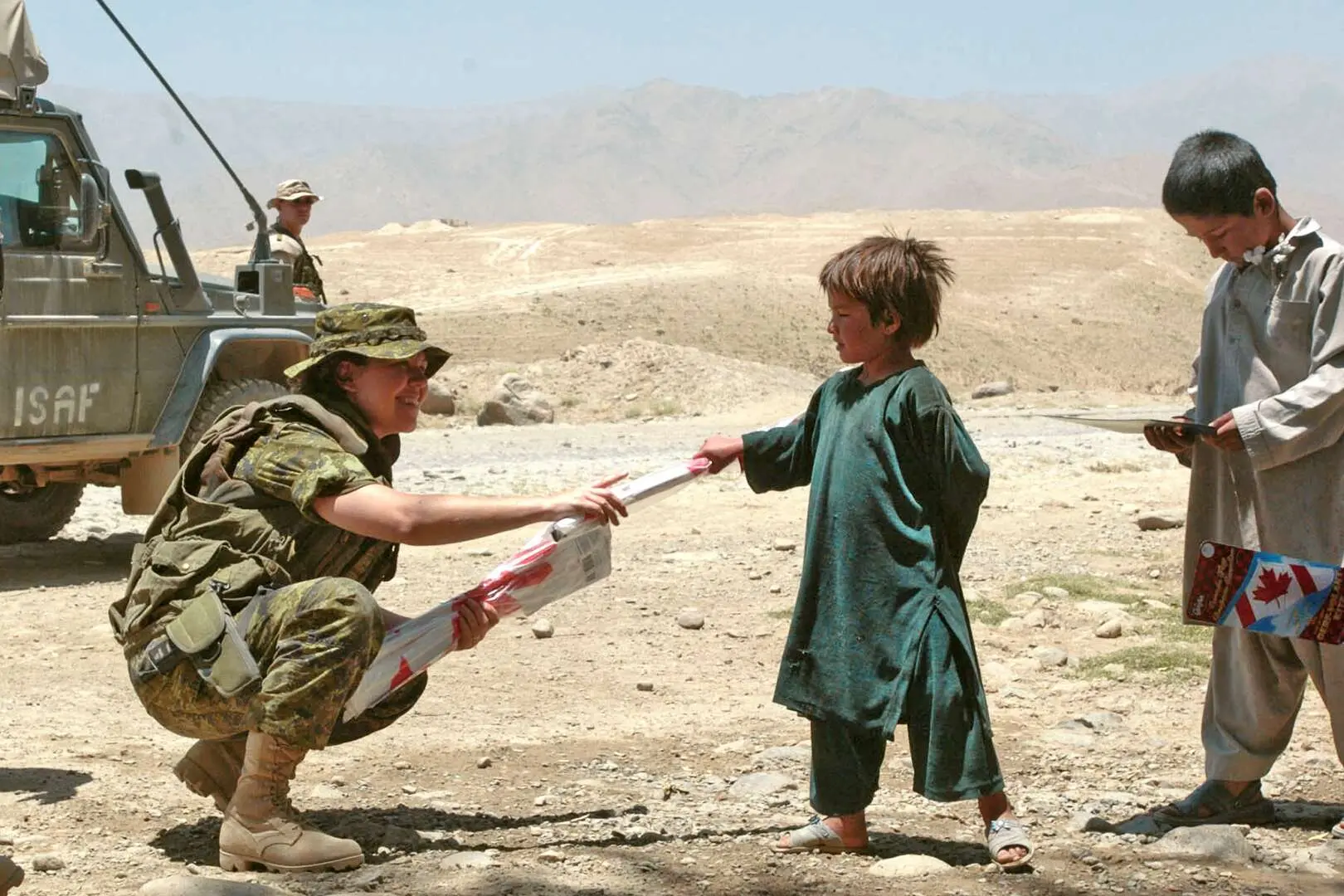 Près d’un véhicule militaire, la capitaine Nancy Pelletier, accroupie, donne un cerf volant aux couleurs du drapeau canadien à un jeune enfant afghan.