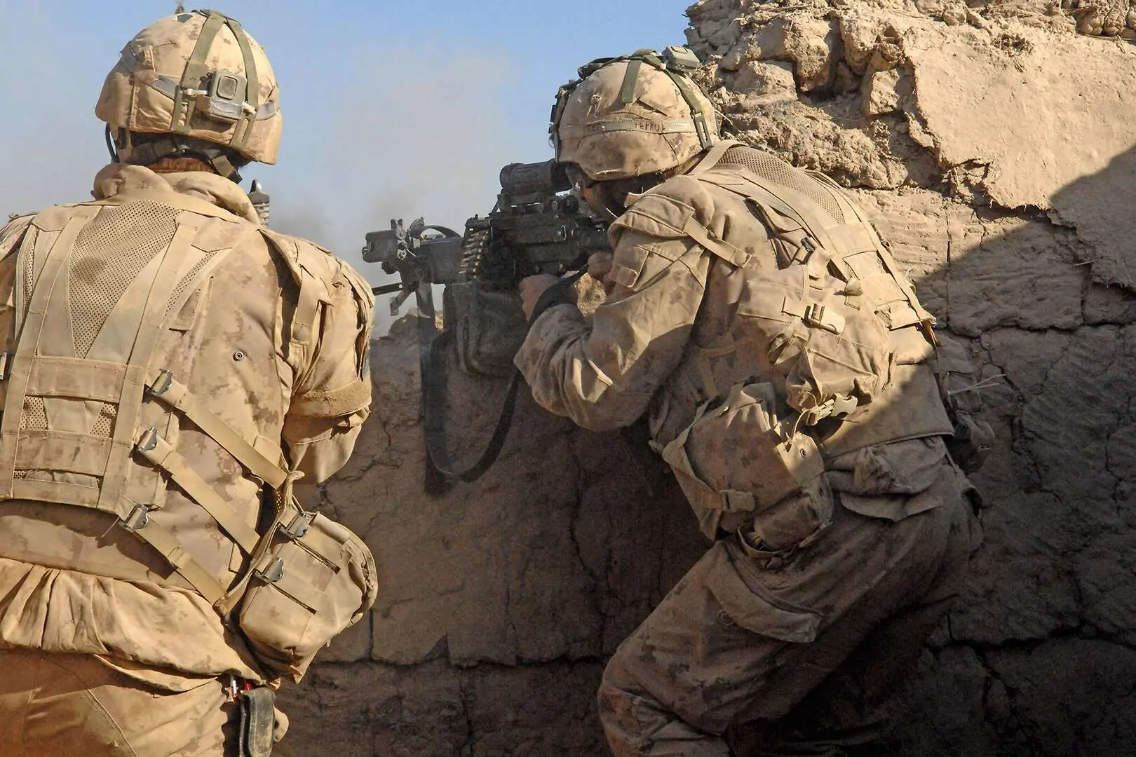 Deux militaires canadiens en habit de camouflage de sable sont dissimulés de dos derrière un muret de pierre. Le soldat se tenant à droite fait feu vers des ennemis avec sa mitrailleuse appuyée sur la pierre.