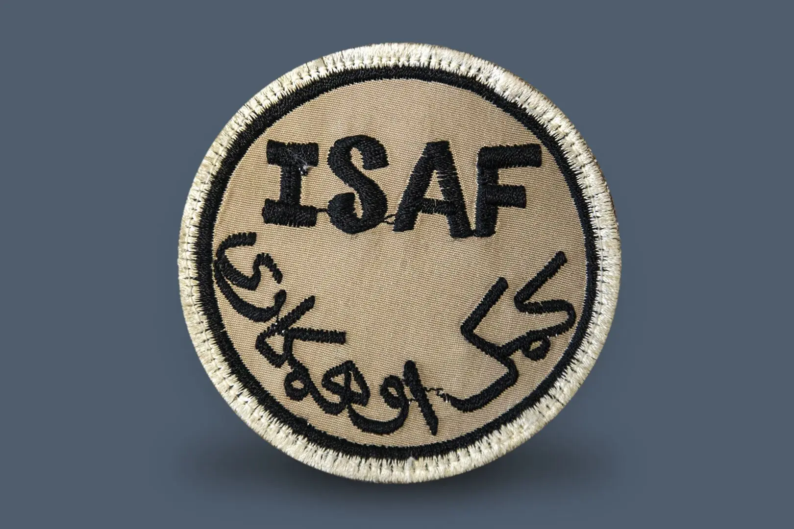 Écusson rond en tissu beige comportant la mention écrite ISAF, acronyme de la Force internationale d’assistance à la sécurité.