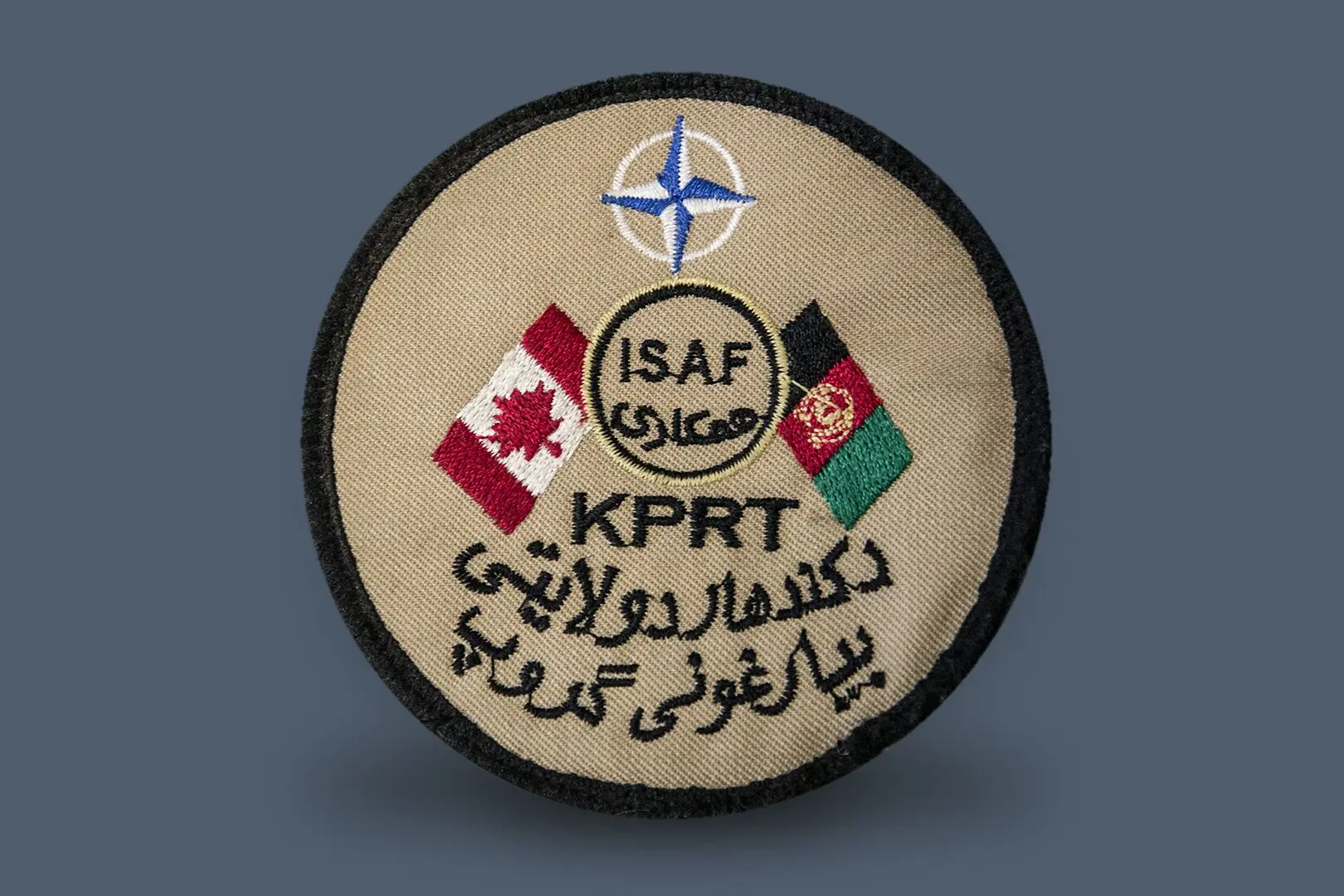 Écusson rond en tissu beige décoré d’une rose des vents, d’un drapeau canadien, d’un drapeau afghan et des mentions écrites ISAF et KPRT au centre de l’écusson. ISAF est l’acronyme de Force internationale d’assistance à la sécurité, tandis que KPRT signifie Équipe provinciale de reconstruction à Kandahar.