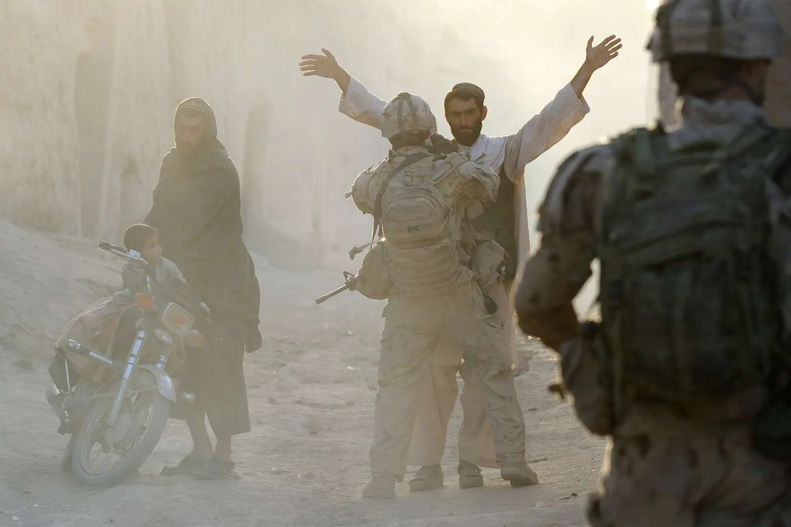 Lors d’un contrôle dans une rue envahie de poussière de sable, un militaire canadien fouille un civil afghan sous l’œil vigilant de son frère d’armes. Sur le côté, un autre civil afghan attend avec un jeune garçon près d’une moto. Le mot ATHENA apparaît en gros au centre de l’image.