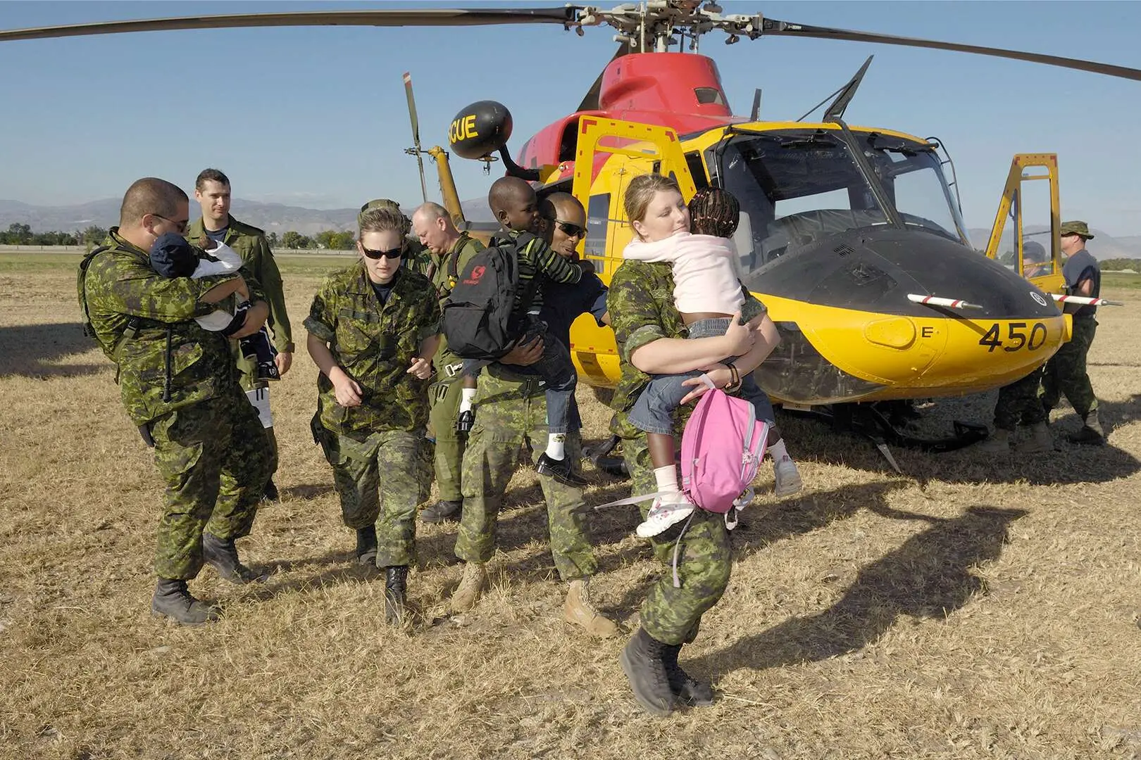 Quatre militaires en tenue de combat portent dans leurs bras trois jeunes Haïtiens près d’un hélicoptère Griffon jaune et rouge de l’Aviation royale canadienne. L’appareil est posé au sol dans un champ. Quatre membres de son équipage circulent autour de celui-ci.