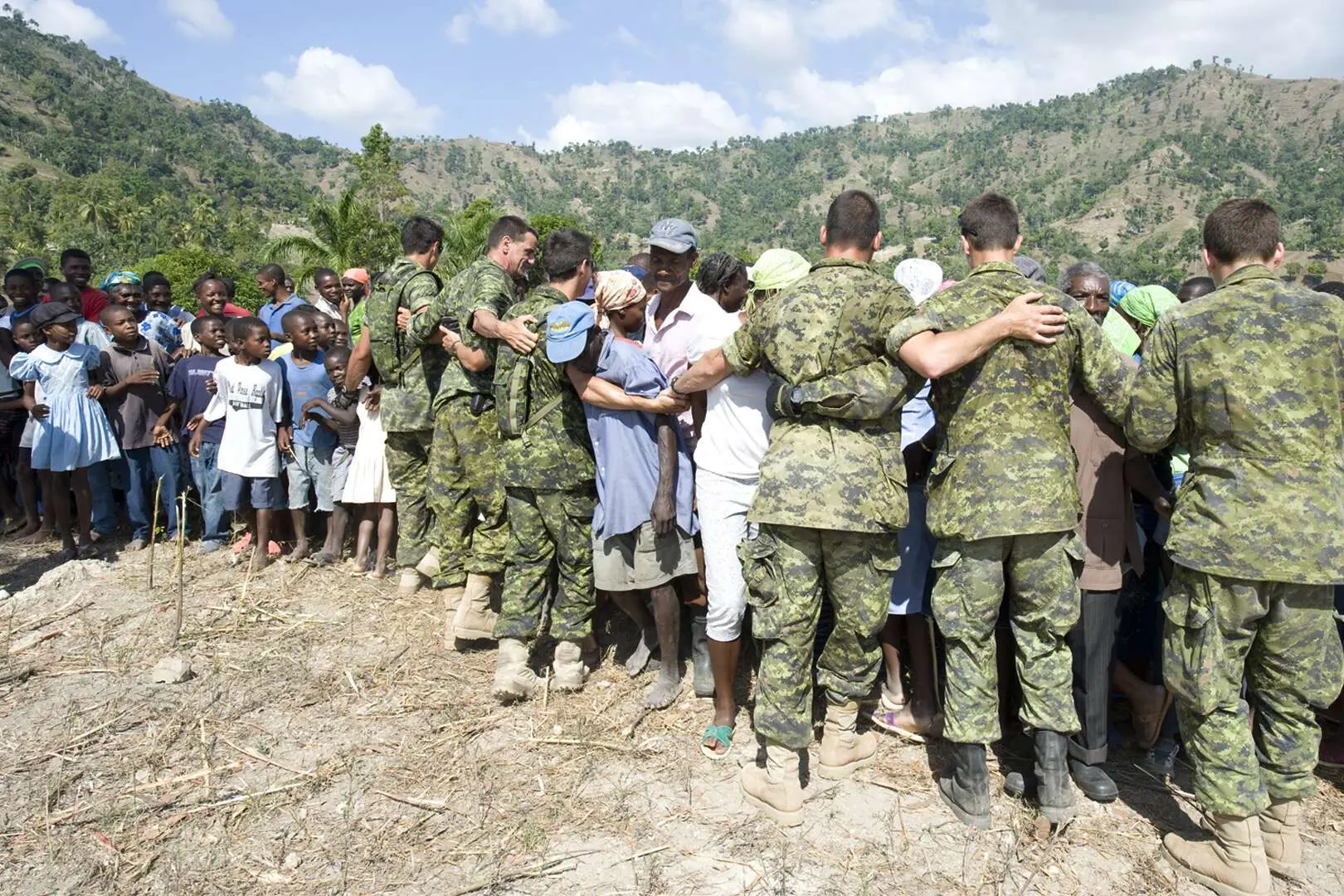 Six militaires canadiens portant l’uniforme de combat de camouflage vert s’unissent avec des Haïtiens pour former une barrière humaine avec en toile de fond une montagne et un ciel bleu parsemé de nuages. Parmi les Haïtiens, on trouve des hommes, des femmes et des enfants. 