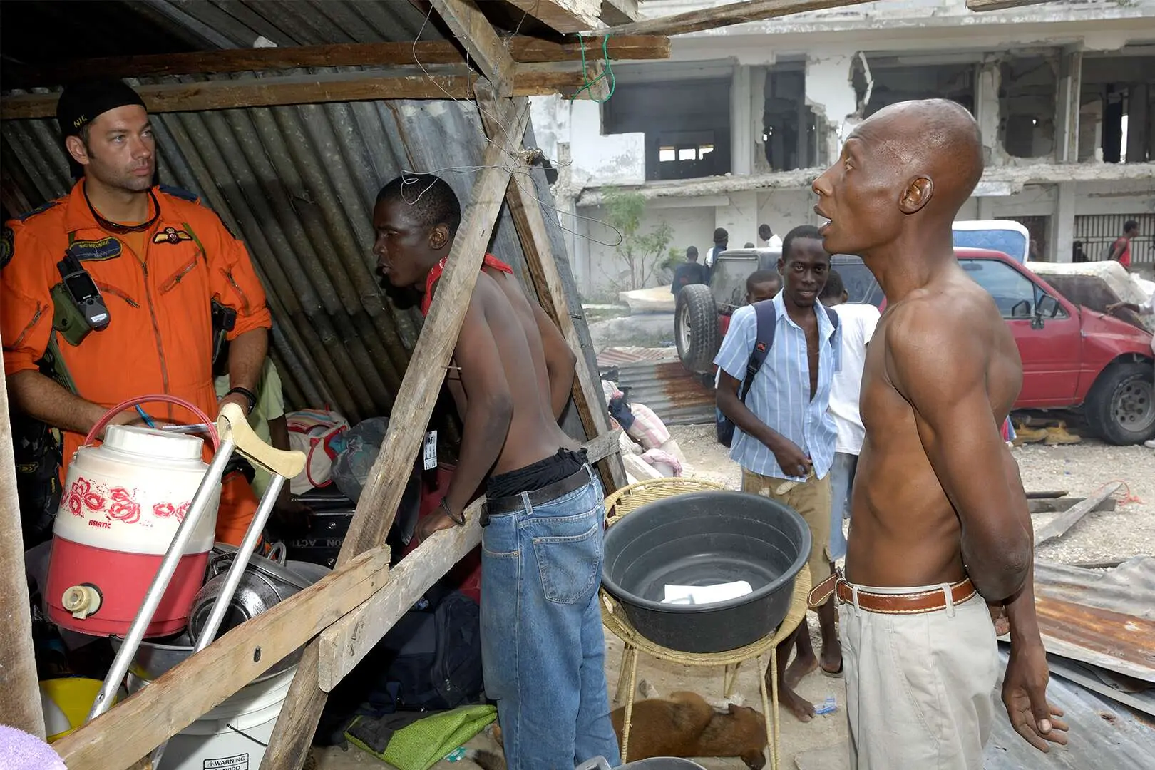 Sous un abri en tôle situé au milieu des ruines de bâtiments détruits suite au tremblement de terre, un militaire à l’air déterminé et vêtu d’un uniforme orange tient entre ses mains un crayon et du papier. Il discute avec deux Haïtiens, torse nu.