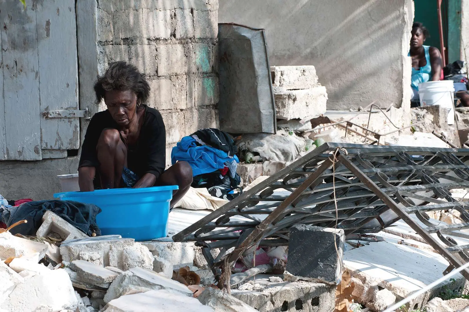 Une Haïtienne qui semble démunie est assise à travers des débris de ciment et de métaux pour faire sa lessive dans un bac bleu.