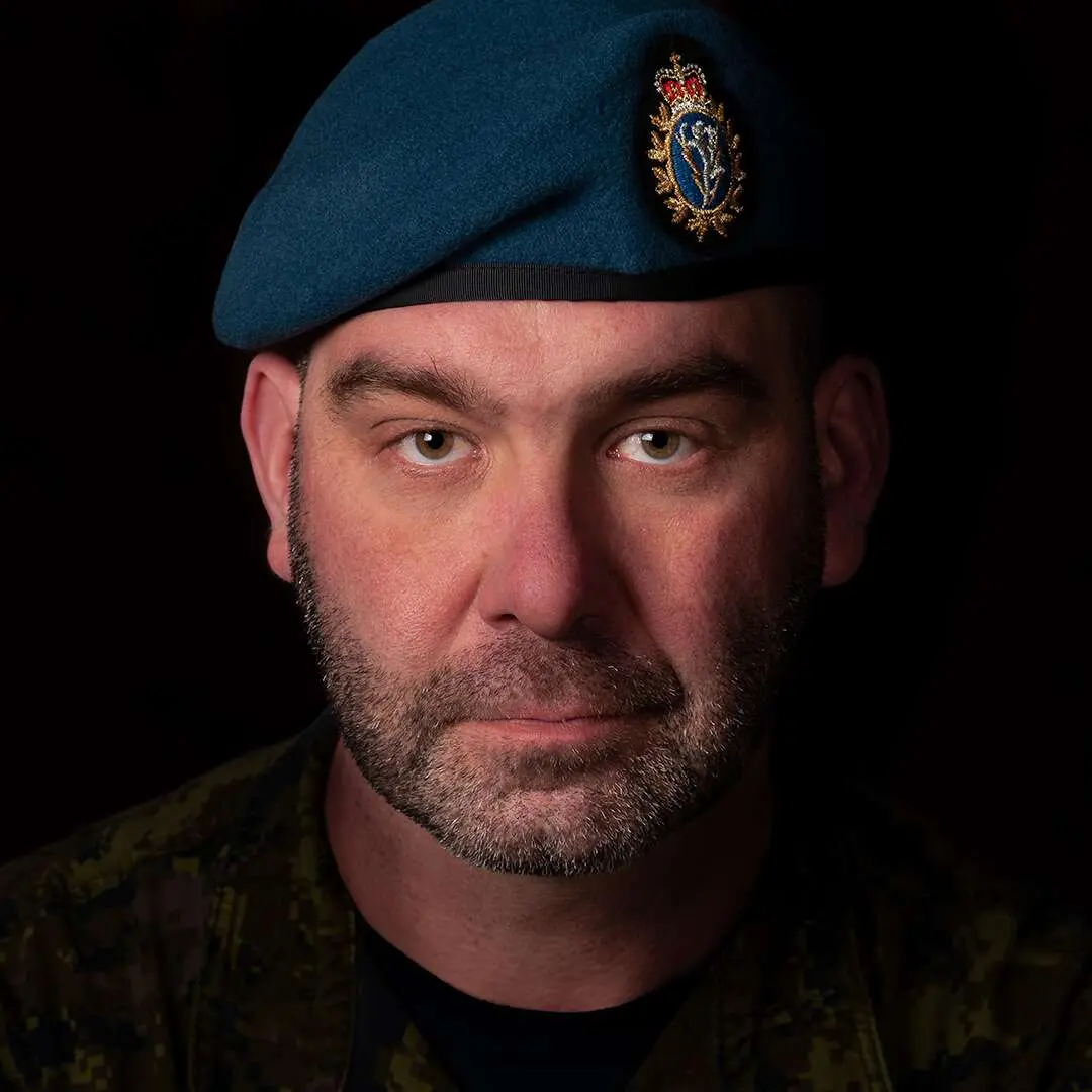 Sur un fond noir, l’adjudant Sébastien Turgeon affiche un air impassible. Il possède une légère barbe foncée et porte son béret bleu de l’Aviation royale canadienne.