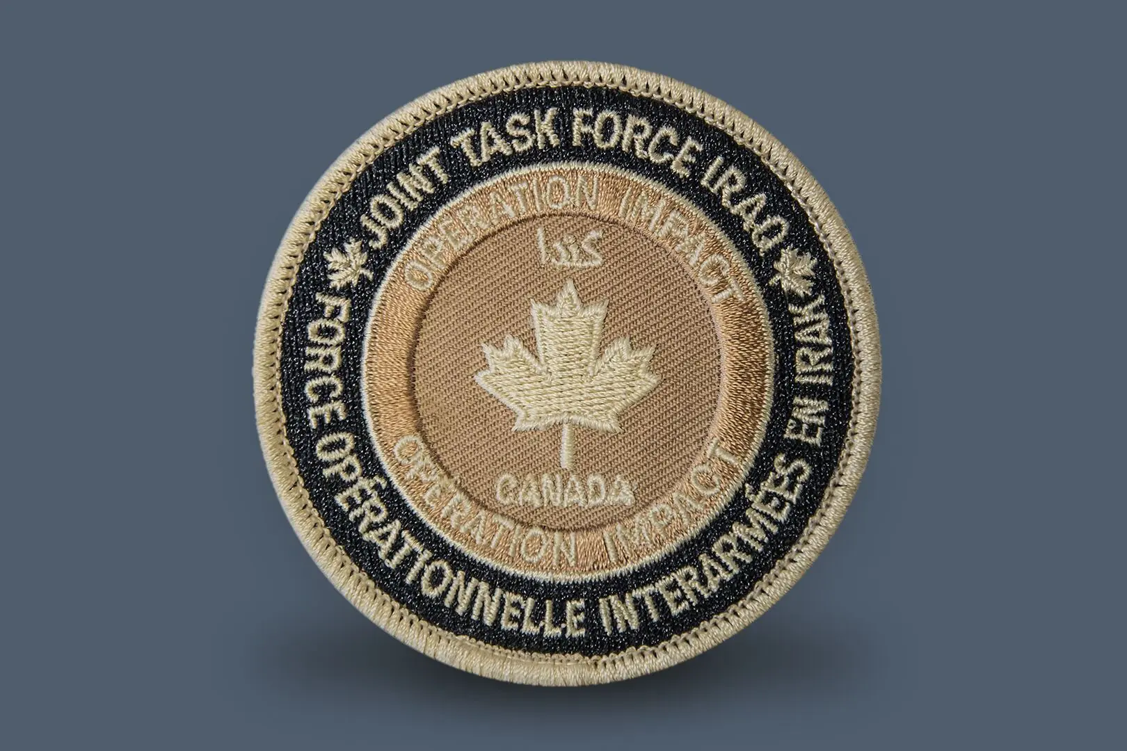 Écusson circulaire en tissu. Sur le pourtour noir sont inscrits «&nbsp;Joint Task Force Iraq&nbsp;» en haut et «&nbsp;Force opérationnelle interarmées en Irak&nbsp;» en bas. Au centre, une feuille d’érable sur fond beige est entourée des mots «&nbsp;Canada&nbsp;» et «&nbsp;Opération IMPACT&nbsp;».