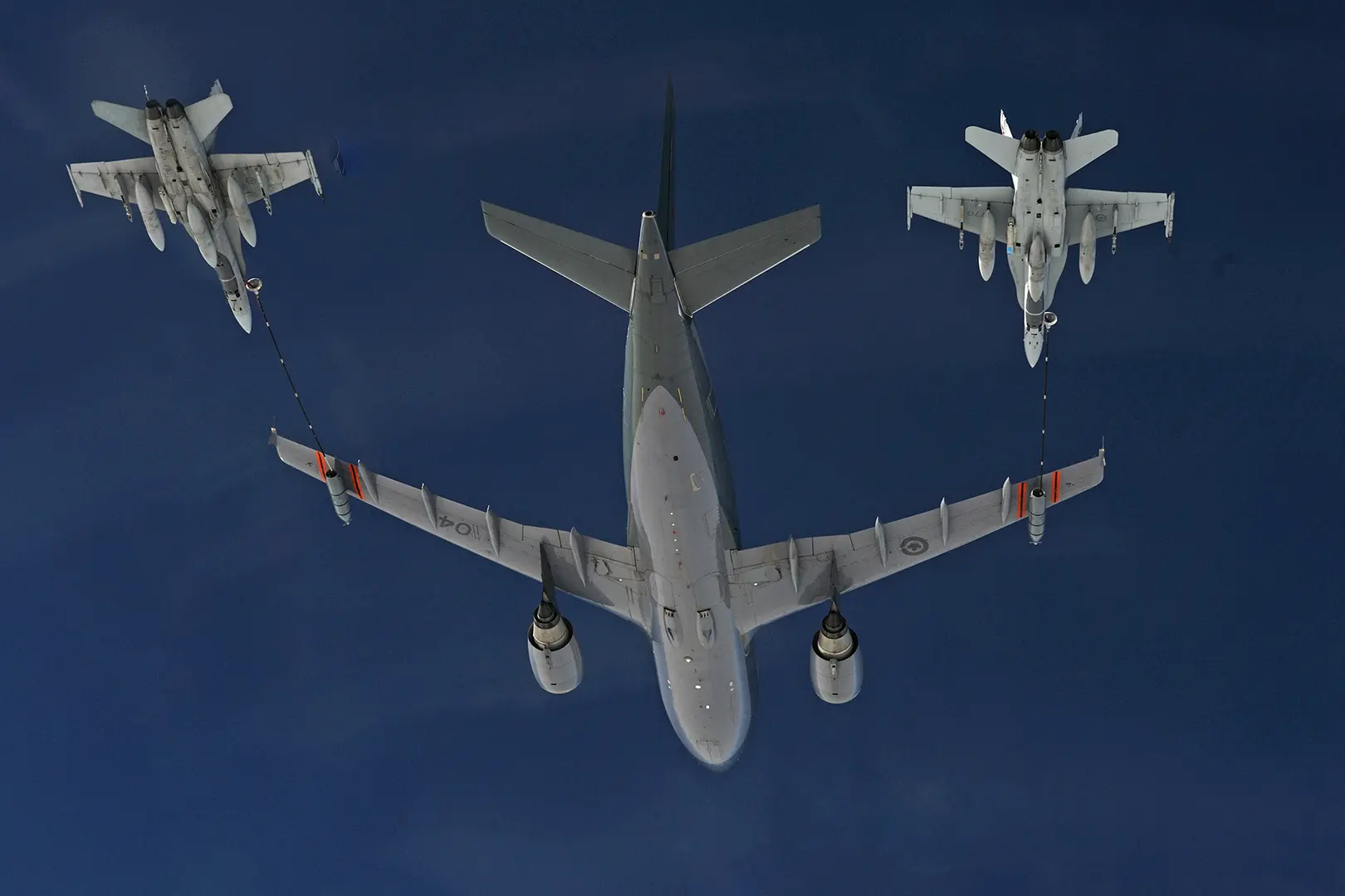 Trois aéronefs sont vus d’en dessous. Un CC-150 Polaris gris ravitaille deux chasseurs CF-18 Hornet situés derrière lui. On peut apercevoir les deux tuyaux de ravitaillement qui rejoignent les deux chasseurs dans un ciel bleu sans nuage.