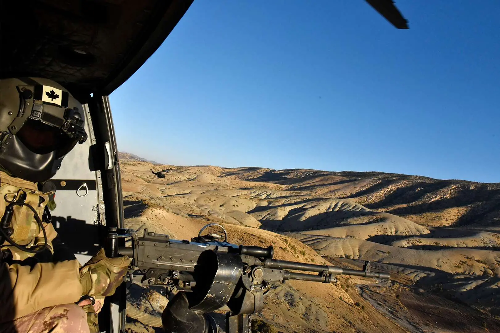 Un mitrailleur est assis dans un hélicoptère, porte ouverte. Il est habillé de son uniforme camouflage beige et d’un casque gris à visière noire. Le militaire tient entre ses mains une mitrailleuse installée sur un trépied. Au sol, on peut voir des collines de sable dépourvues de végétation s’étendre à l’infini sous un ciel bleu. 