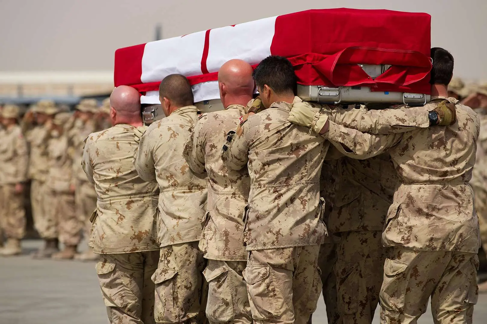 Des militaires canadiens portent un cercueil. Les militaires sont revêtus de leur uniforme de camouflage beige et marchent de dos avec le cercueil sur l’épaule. Celui-ci est recouvert d’un drapeau canadien. 