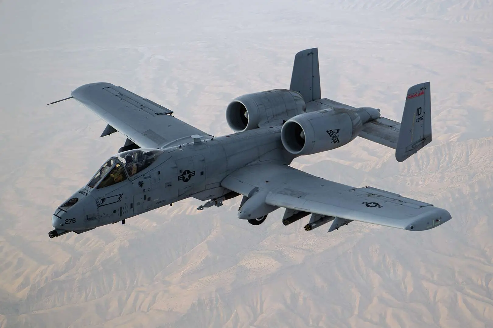Un avion A-10 Thunderbolt gris et identifié par le logo de l’US Air Force survole une zone désertique avec des collines de sable. Le A-10 possède deux turboréacteurs surélevés. On peut voir cinq missiles sous ses ailes et un canon au-devant de l’appareil. 