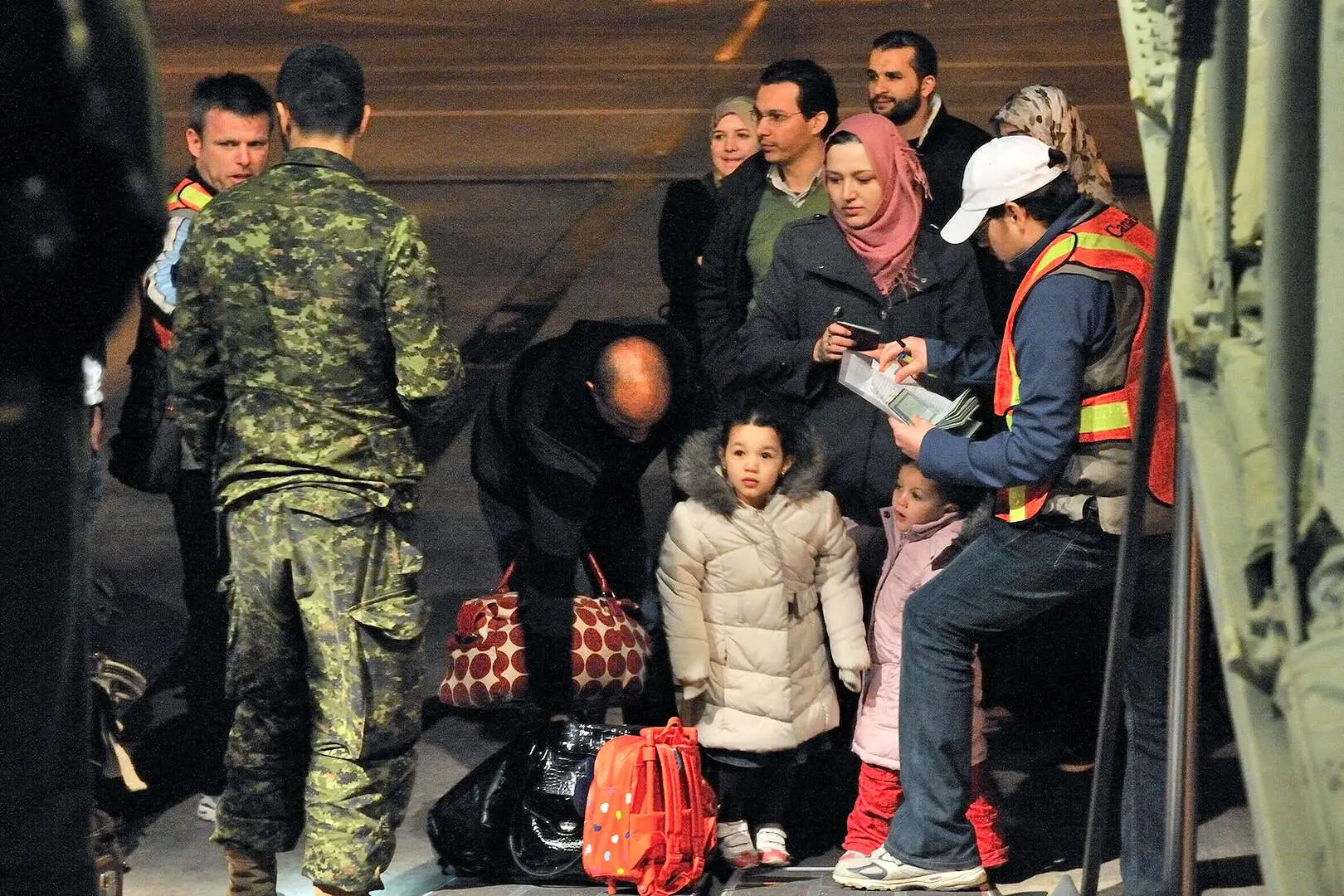 À l’entrée de l’avion, deux officiers de sécurité et un militaire canadien réalisent une opération de contrôle auprès de huit civils libyens. Au premier plan, deux enfants habillés de gros manteaux patientent. Derrière eux se tiennent trois femmes et trois hommes équipés de sacs de voyage.
