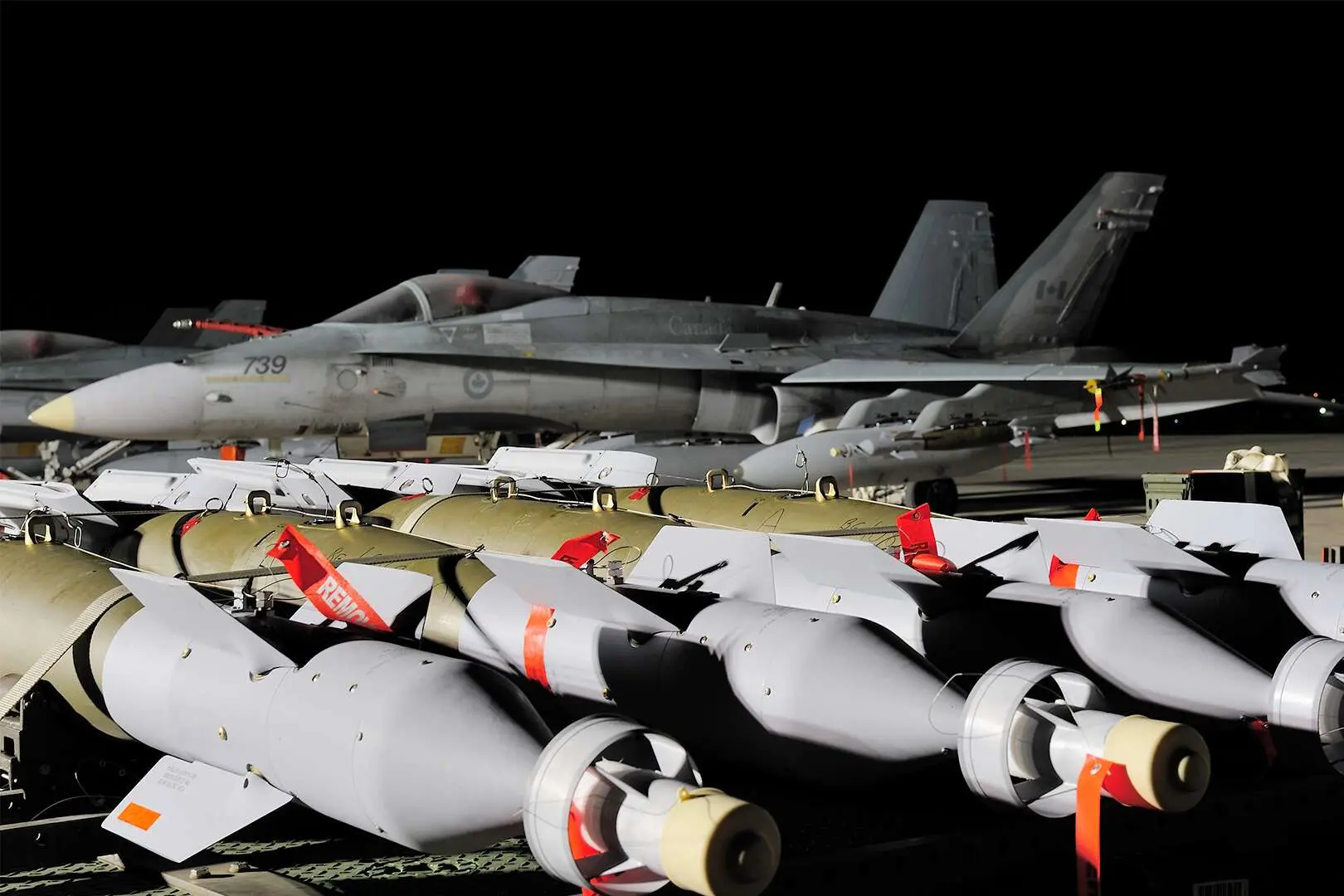 Sur un fond noir, un avion CF-18 canadien de couleur grise est immobile avec son flanc gauche à découvert. En avant-plan, trois missiles gris et dorés sont visibles.