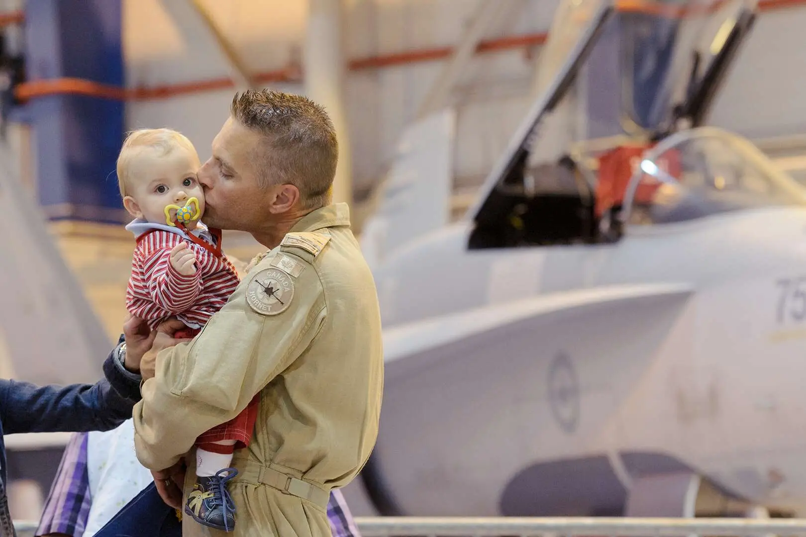 Le lieutenant-colonel Sylvain Ménard, revêtu de son uniforme de pilote, embrasse son bébé, un jeune garçon blondinet, qu’il tient dans ses bras. Derrière, on peut apercevoir un CF-18 gris à l’intérieur d’un hangar.
