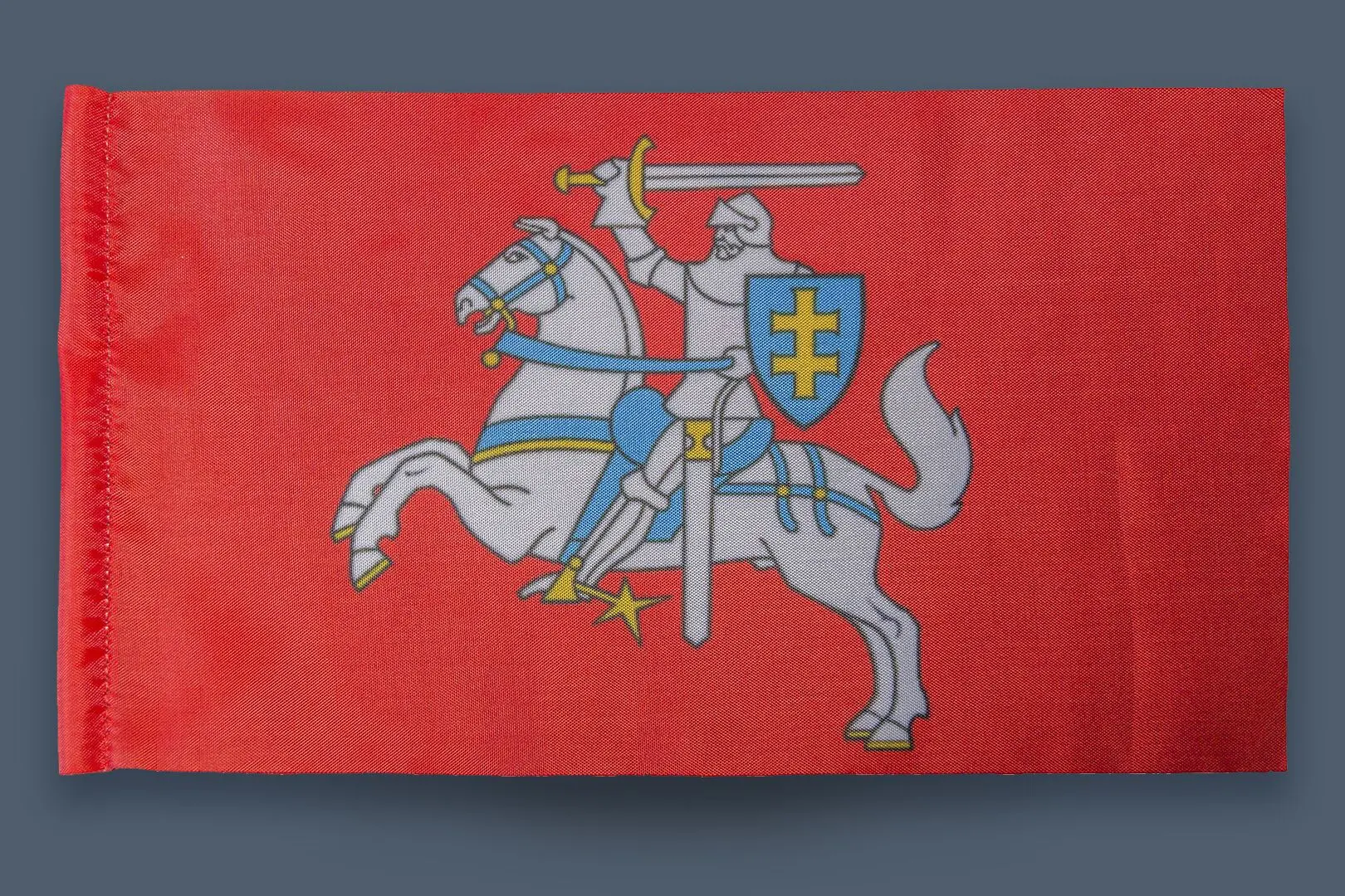 Drapeau rouge décoré en son centre d’un chevalier gris assis sur un cheval. Le personnage porte un bouclier au bras gauche et brandit une épée avec sa main droite tout en tirant sur la bride de sa monture.