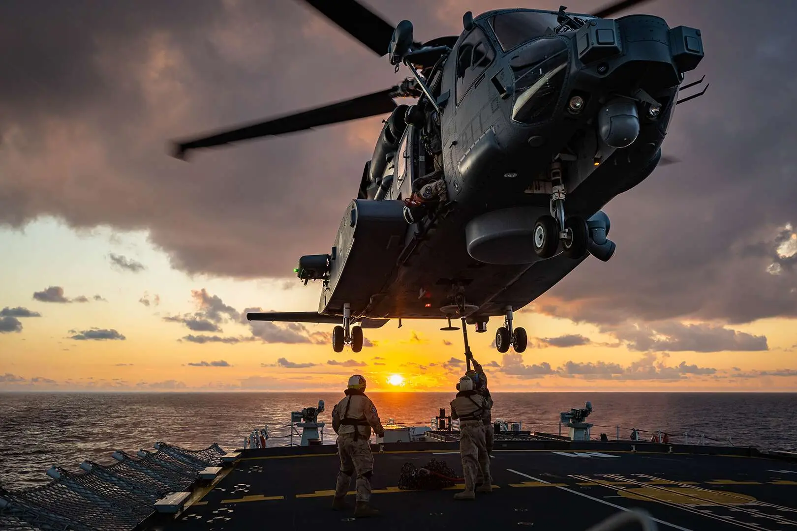 Un hélicoptère est stabilisé au-dessus du pont d’un navire. Trois militaires sont en position sous l’appareil afin de réaliser un exercice de treuillage. En toile de fond, on peut voir le ciel parsemé de nuages et le soleil orangé qui s’apprête à plonger dans la mer aux reflets gris-mauve.