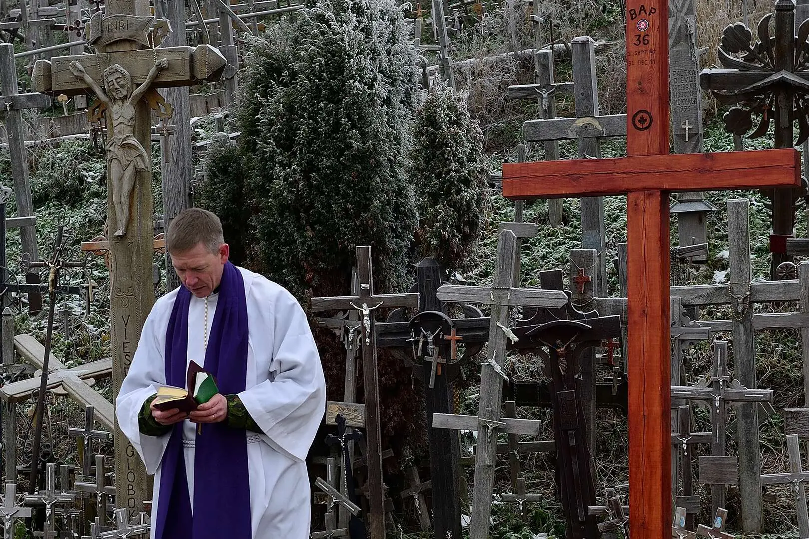 Revêtu de son habit blanc, l’aumônier tient deux petits livres entre ses mains. En arrière-plan se dresse une colline ornée de dizaines de croix. 
